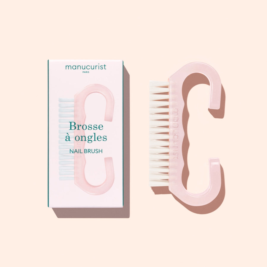 A translucent pink Nail Brush with vegan bristles and white tips from Manucurist-us rests next to its box, which displays an image of the brush and the words “Manucurist Paris, Brosse à ongles, NAIL BRUSH,” set against a pale background—ideal for enhancing your manicure routine.