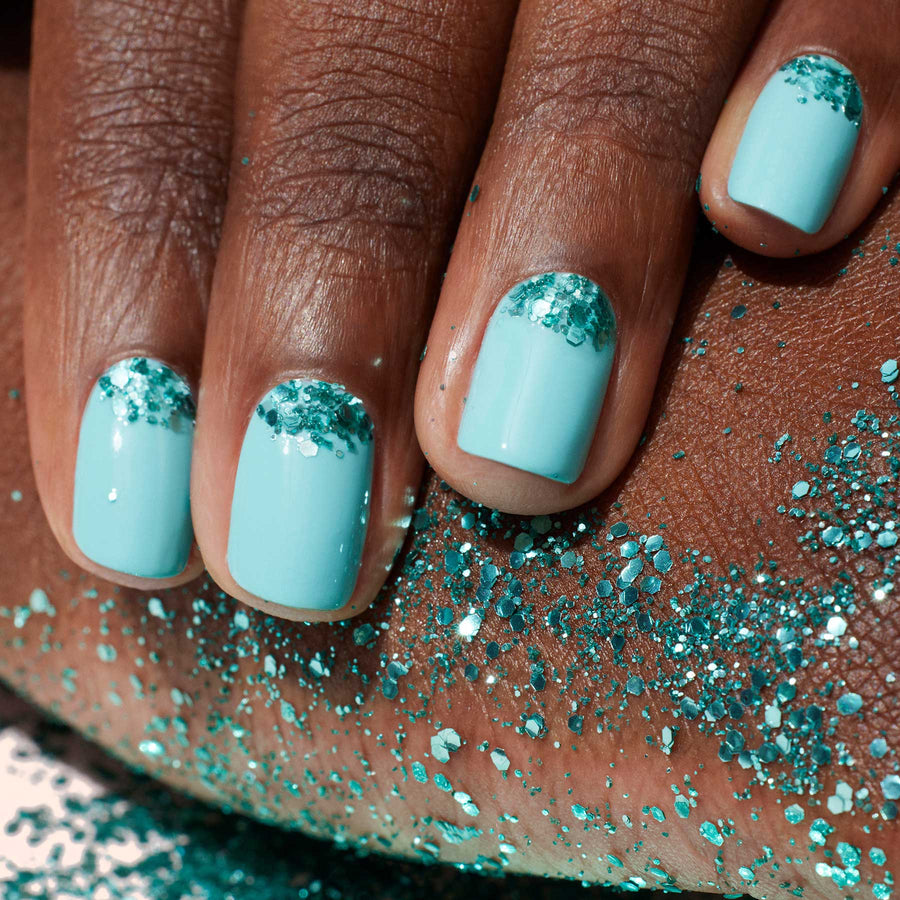 A bottle of Manucurist Underwater Glitter nail polish, a small open jar of biodegradable glitter, and a wooden manicure tool are arranged on a coiled white rope with navy accents.