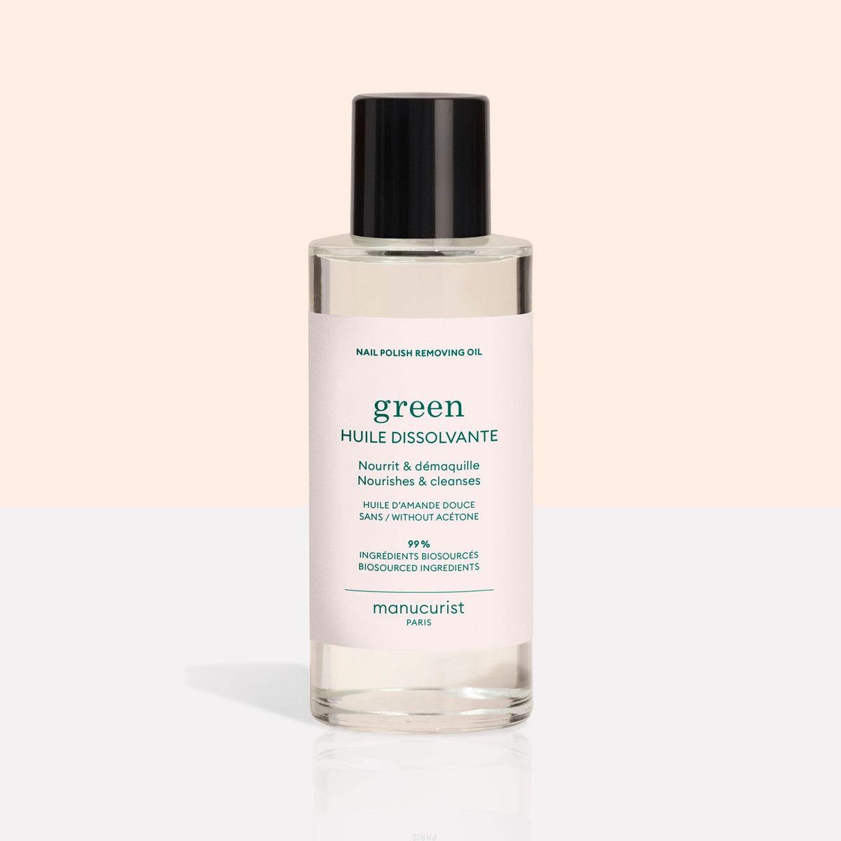 A clear bottle with a black cap contains Manucurist Nail Polish Removing Oil, a green, plant-based formula. The bottle features a light pink label with green text on a beige and white background.