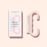 A translucent pink Nail Brush with vegan bristles and white tips from Manucurist-us rests next to its box, which displays an image of the brush and the words “Manucurist Paris, Brosse à ongles, NAIL BRUSH,” set against a pale background—ideal for enhancing your manicure routine.