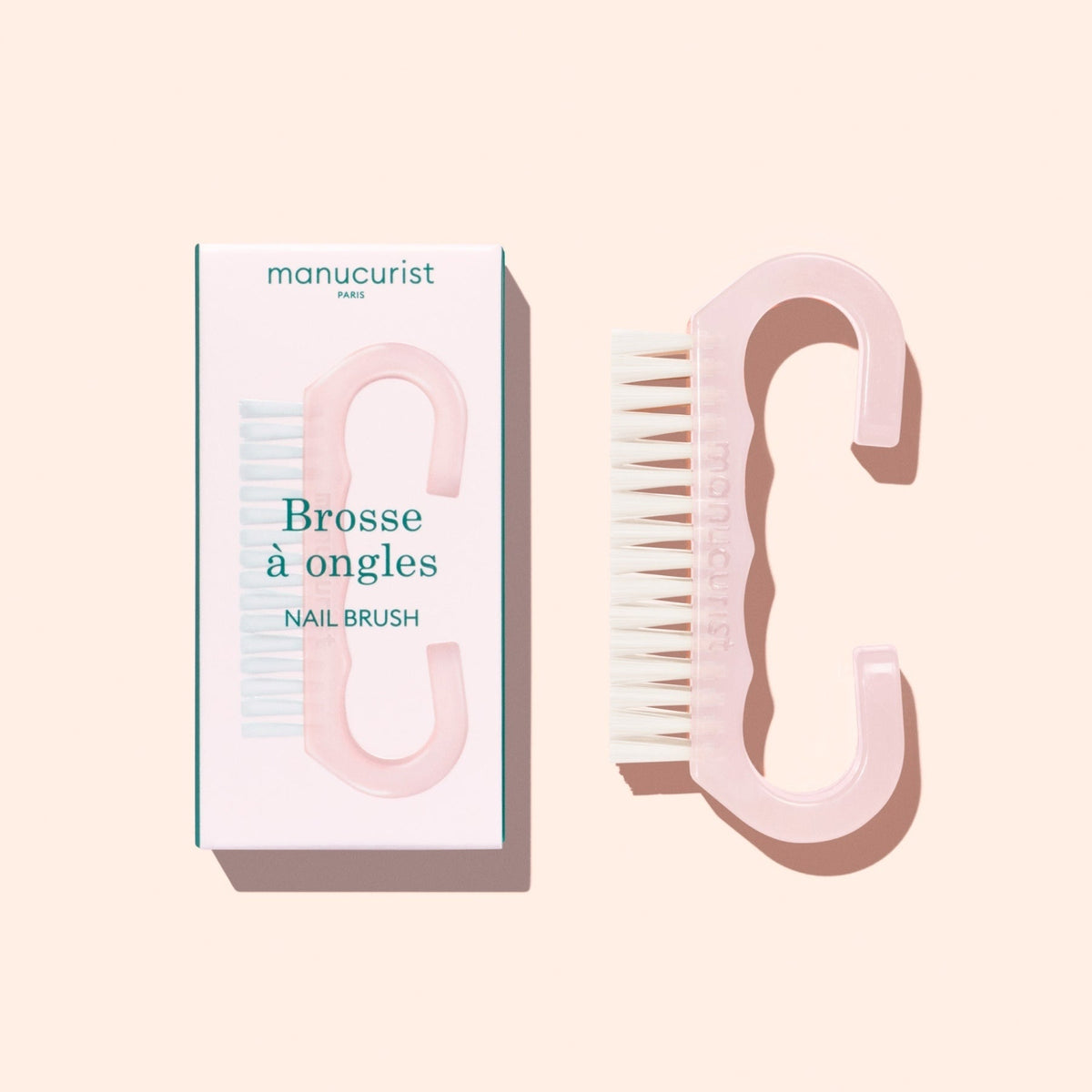 A translucent pink Nail Brush with vegan bristles and white tips from Manucurist-us rests next to its box, which displays an image of the brush and the words “Manucurist Paris, Brosse à ongles, NAIL BRUSH,” set against a pale background—ideal for enhancing your manicure routine.