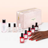 Nine manicure products from Manucurist’s Super Starter Kit—including seven bottles of plant-based nail polish, one remover, and one drops—are arranged above a pink zippered makeup bag labeled “manucurist PARIS” on a light pink background.