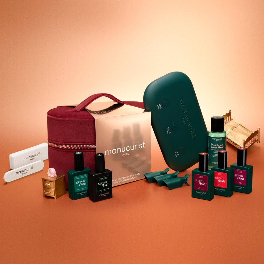 A flat lay of the Manucurist Green Flash™ Starter Kit Gift Set, featuring nail polish bottles, remover, green-capped accessories, a teal nail lamp, nail files, and tweezers—all arranged beside a burgundy zippered pouch on a gradient peach background—perfect for achieving salon-quality manicures at home.