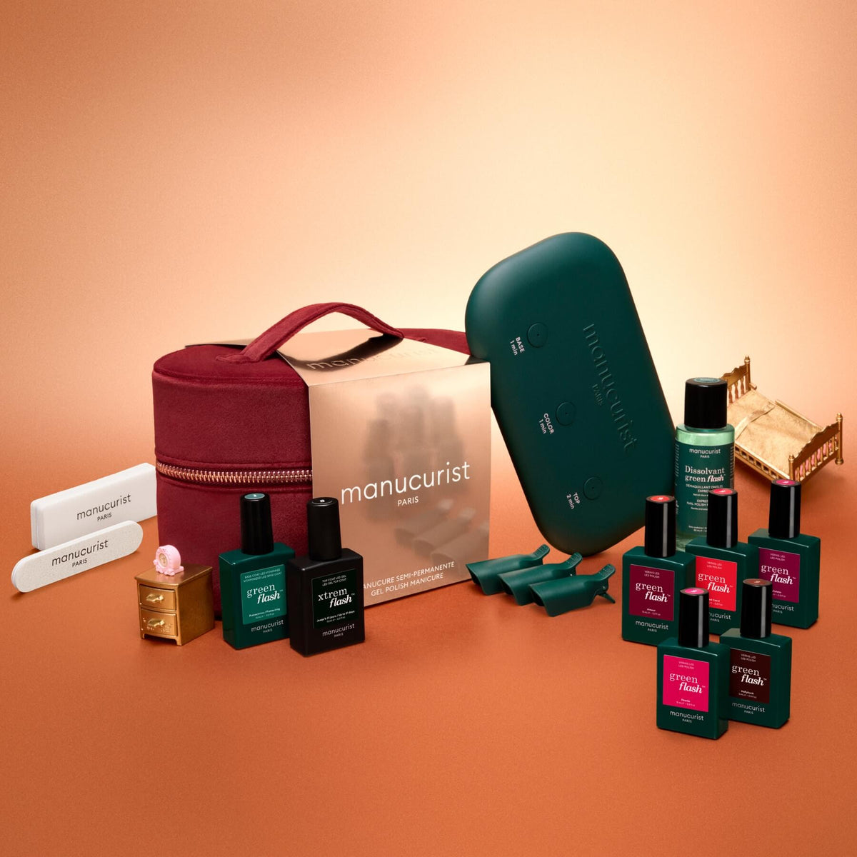 The Manucurist Green Flash™ Super Starter Kit Gift Set features nail polish bottles, remover caps, nail files, a green LED lamp, and manicure tools arranged neatly on an orange background—everything you need for a salon-style manicure at home with long-lasting results.