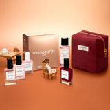 Flat lay of the Manucurist Green™ Discovery Kit Gift Set, featuring a burgundy pouch, a brown Manucurist box, a red gel effect manicure nail polish, and bottles of naturally derived nail polishes and nail care products on a gradient peach background.