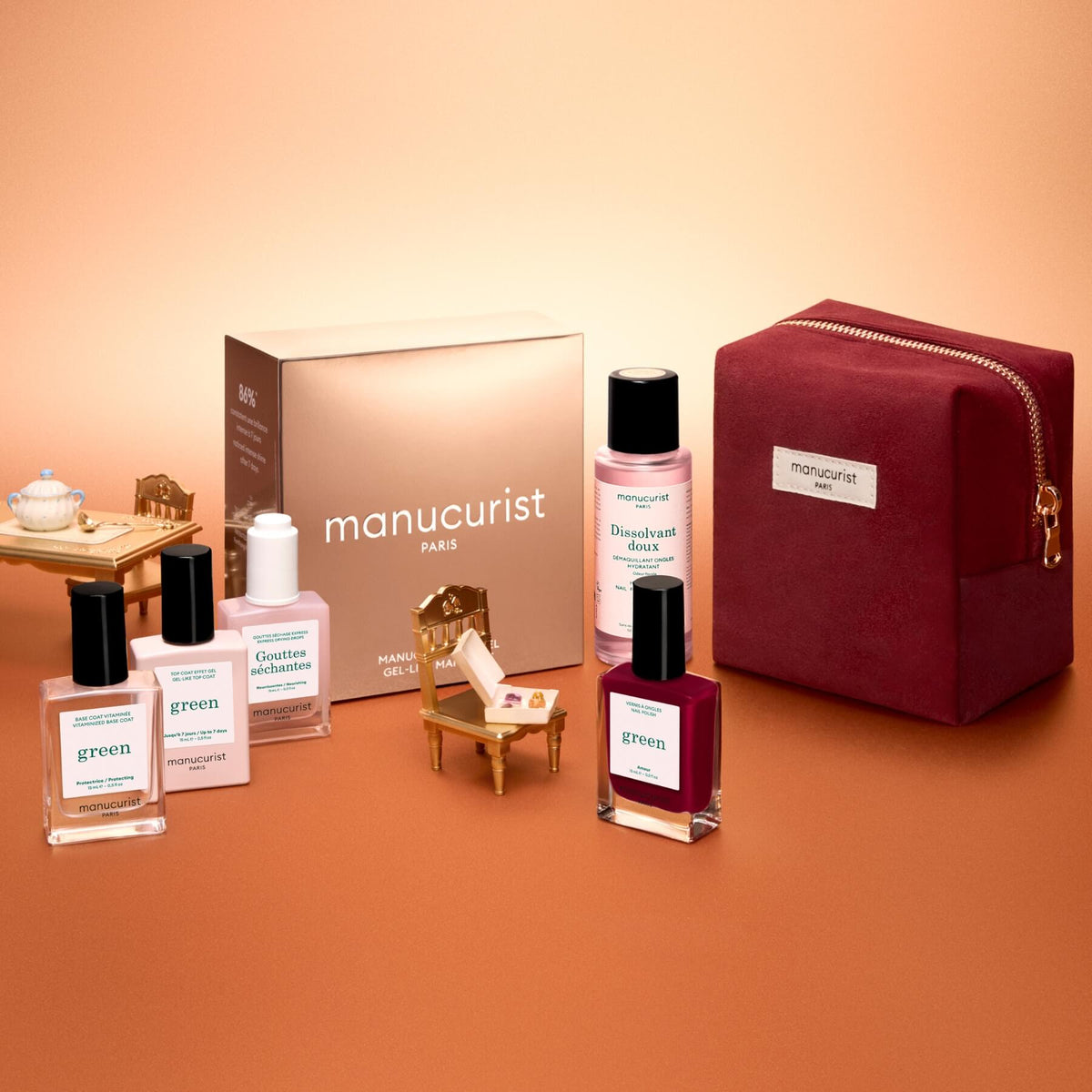 Flat lay of the Manucurist Green™ Discovery Kit Gift Set, featuring a burgundy pouch, a brown Manucurist box, a red gel effect manicure nail polish, and bottles of naturally derived nail polishes and nail care products on a gradient peach background.