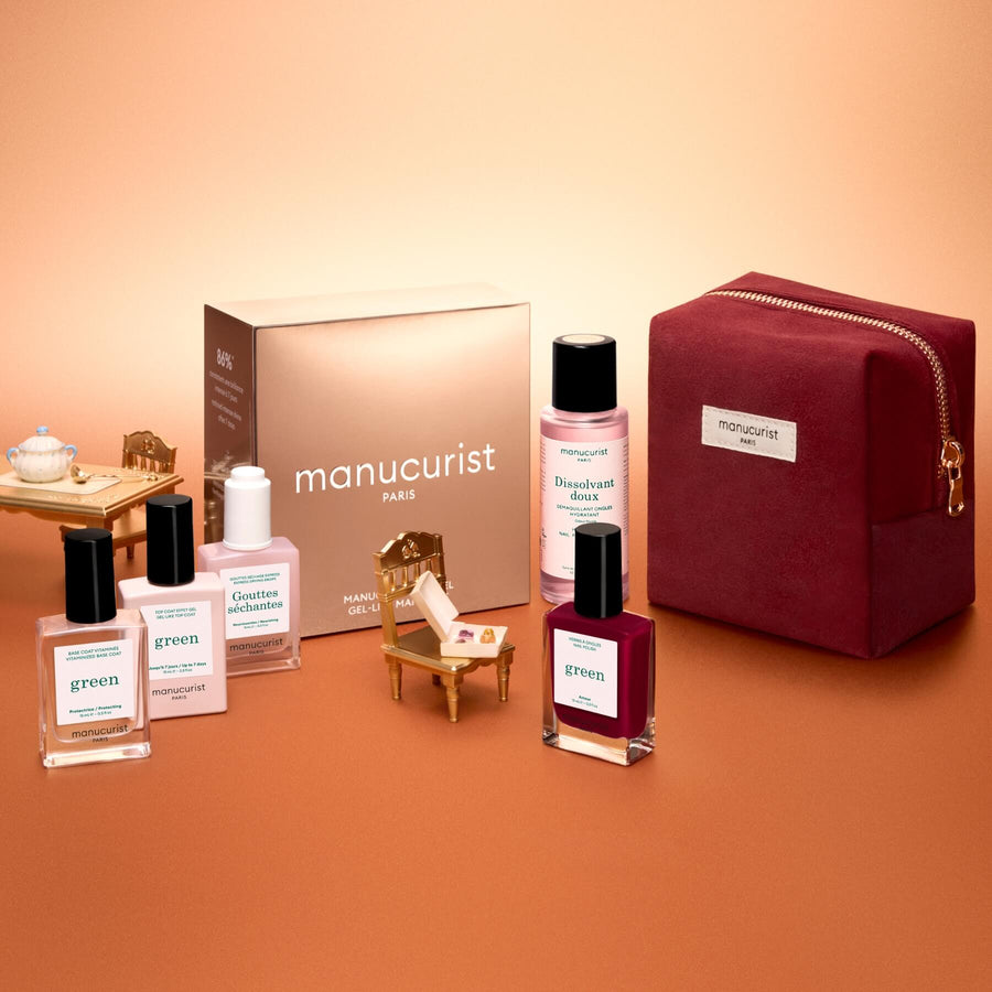 Flat lay of the Manucurist Green™ Discovery Kit Gift Set, featuring a burgundy pouch, a brown Manucurist box, a red gel effect manicure nail polish, and bottles of naturally derived nail polishes and nail care products on a gradient peach background.