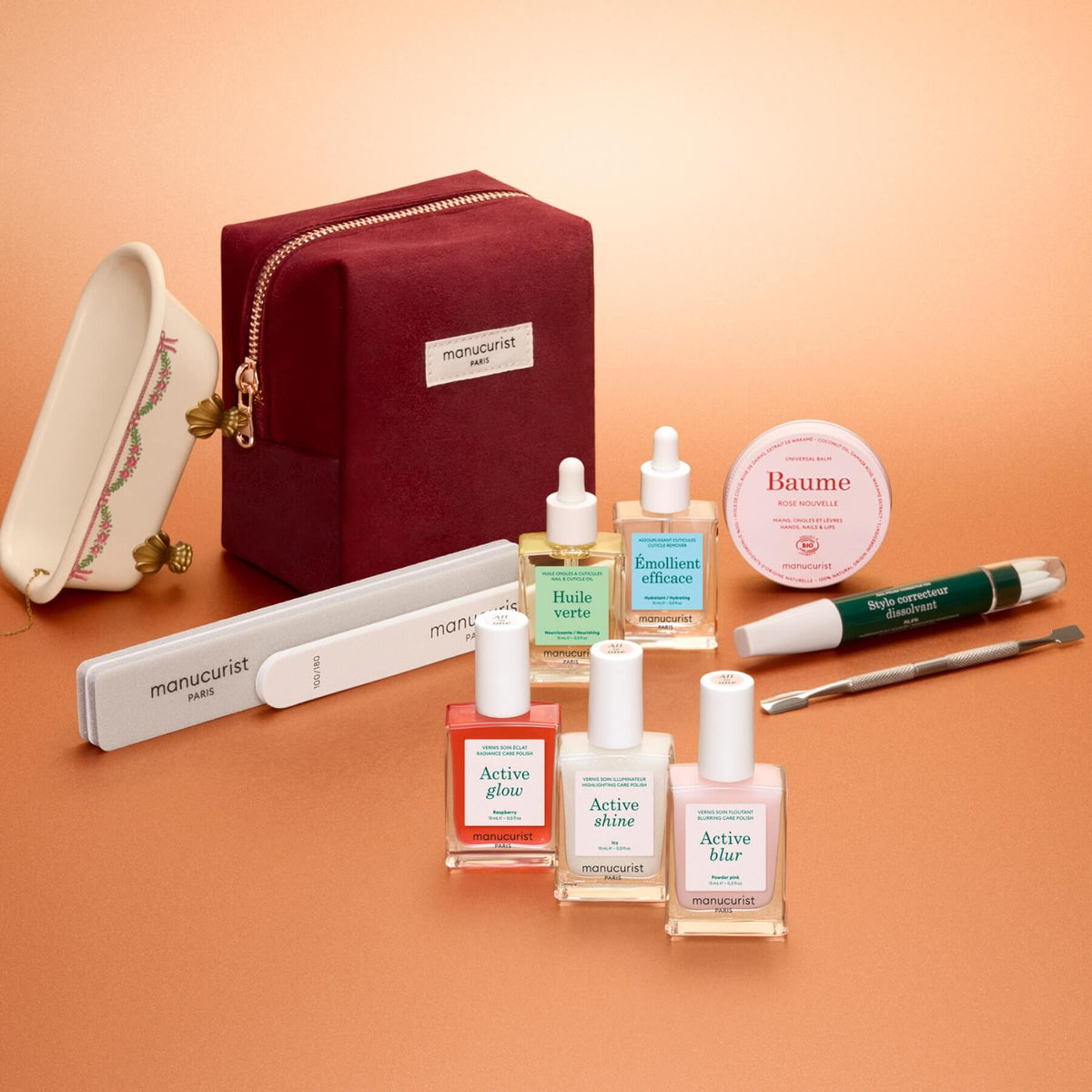 The Manucurist Active™ Ultimate Gift Set, featuring a burgundy pouch, is elegantly displayed on a peach background—surrounded by nail files, a cuticle tool, six nail care bottles, and a round balm tin—making it the perfect upgrade to your manicure routine or a thoughtful holiday gift.