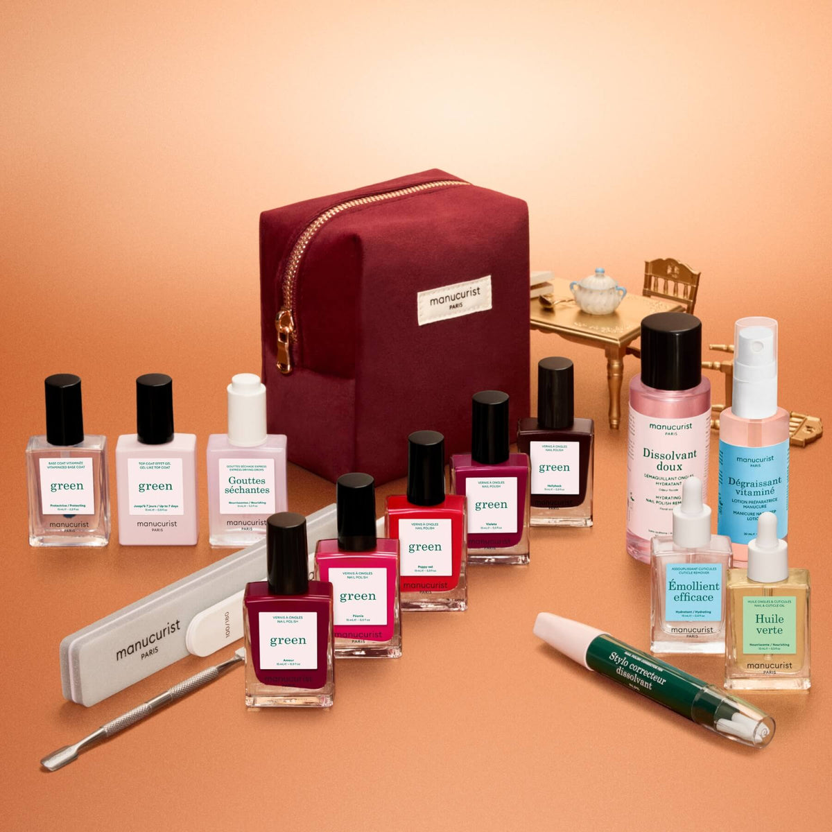 A flat lay of the Manucurist Green™ Super Starter Kit Gift Set, featuring eight naturally derived nail polishes, remover, treatment oils, a file, cuticle pusher, and a burgundy kit bag—everything you need for the ultimate nail prep—beautifully arranged on a peach background.
