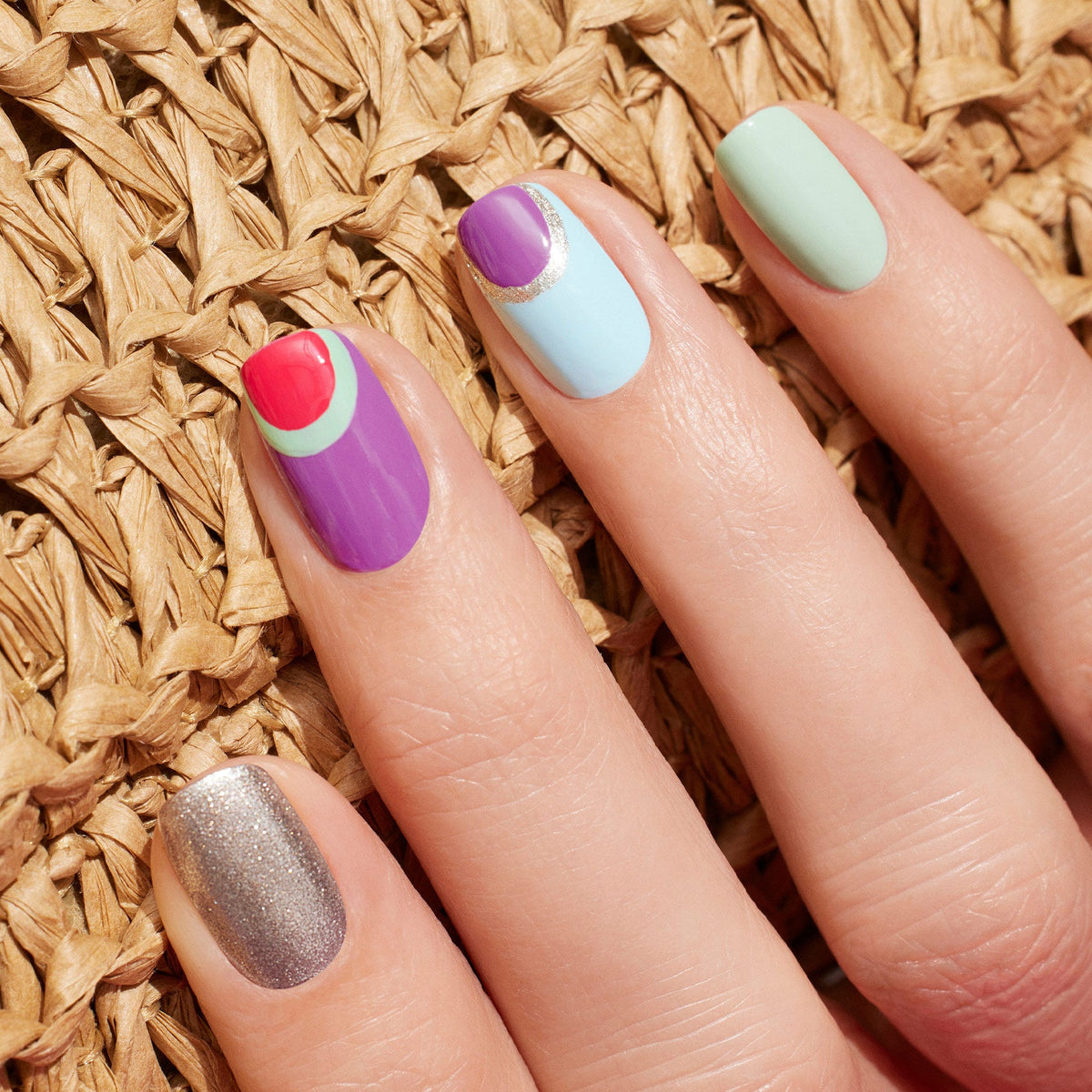A hand with Manucurist's Retro Rainbow nail art holds a box of five Green Flash polishes, neatly arranged on a woven surface alongside a multicolored stone and a sun hat.