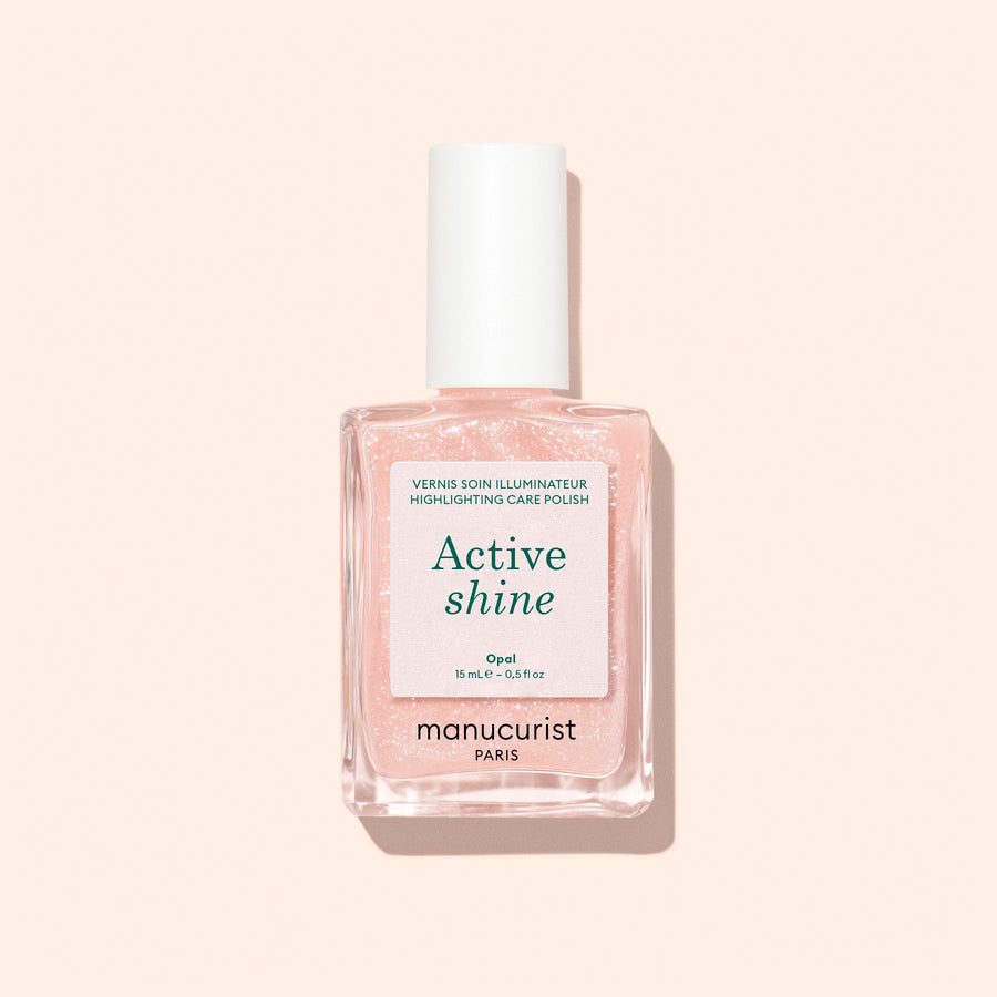 A bottle of Manucurist Active™ Shine Opal plant-based nail polish, featuring a white cap and filled with a light pink, shimmery formula designed to strengthen nails, displayed against a pale beige background.