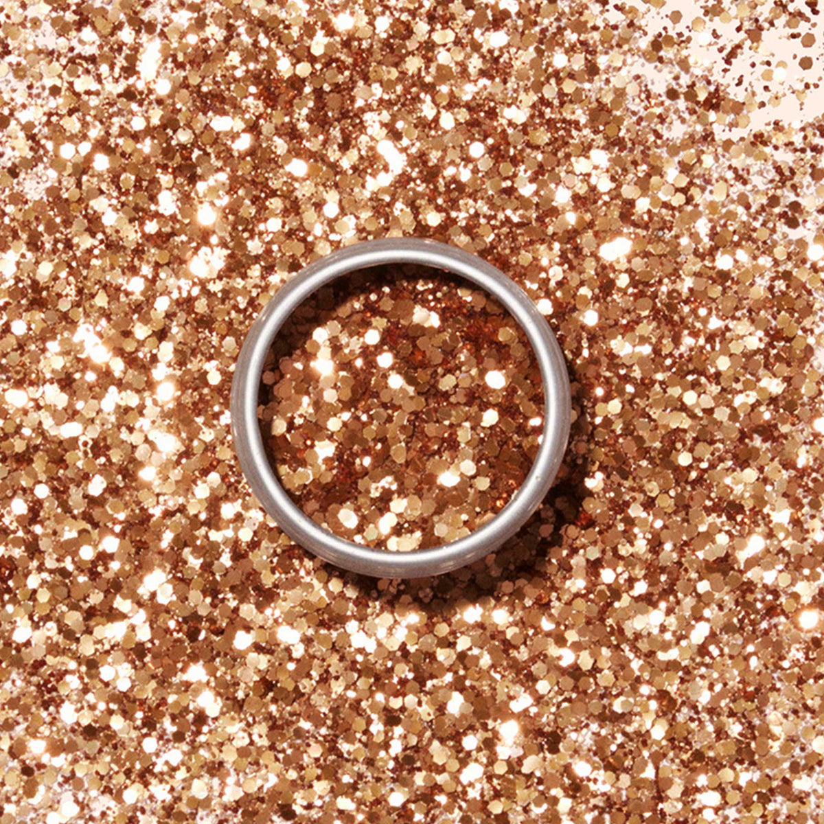 A round metal tin of Manucurist’s Copper Biodegradable Glitter displays gleaming gold and bronze hues against a pale beige background, casting a soft shadow to the left.