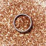 A round metal tin of Manucurist’s Copper Biodegradable Glitter displays gleaming gold and bronze hues against a pale beige background, casting a soft shadow to the left.