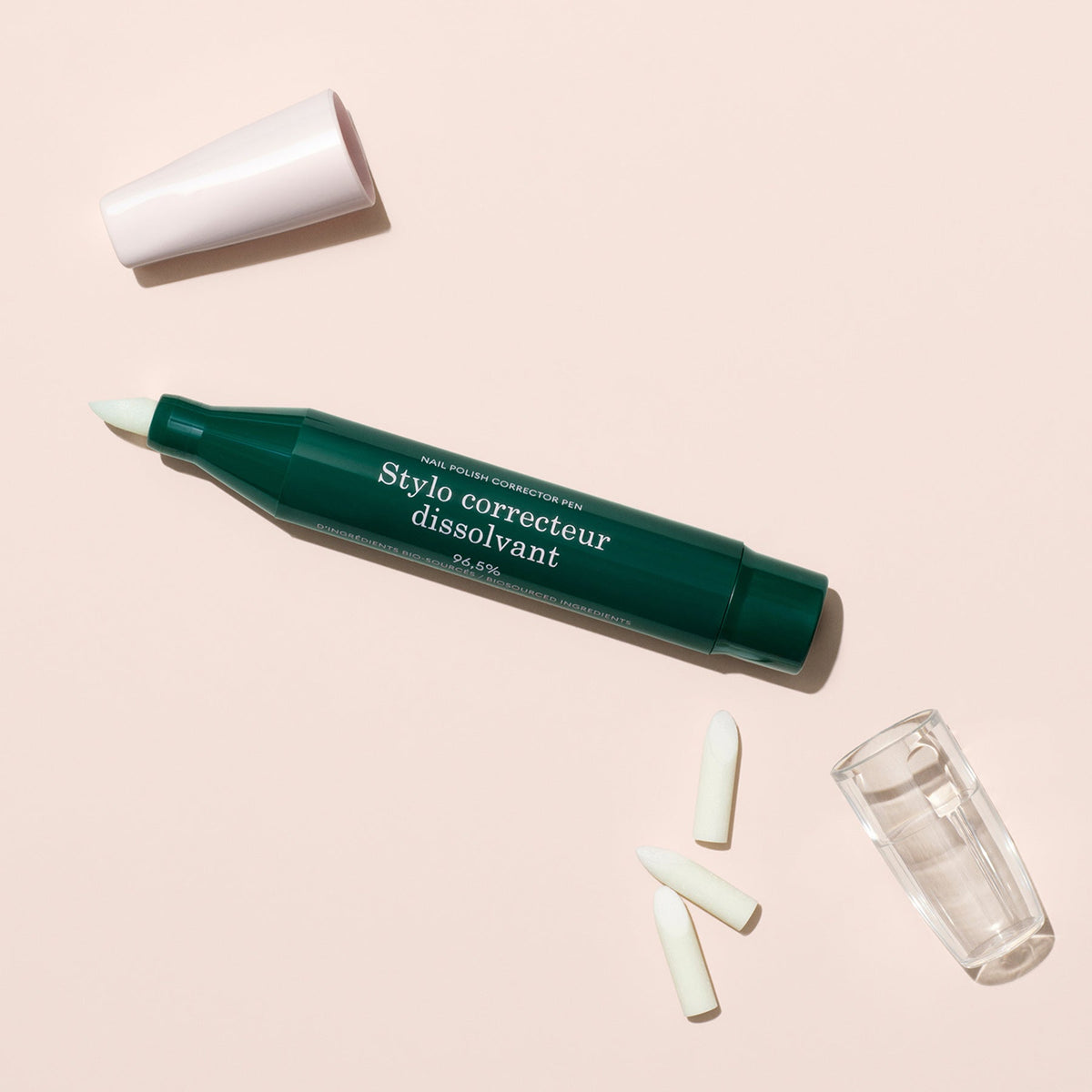 A Manucurist-test + Corrector Pen, green and white with "Stylo correcteur dissolvant" on it, lies diagonally on a light beige background. The cap covers one end, while a clear cover reveals its fine tip for precise gel polish correction.