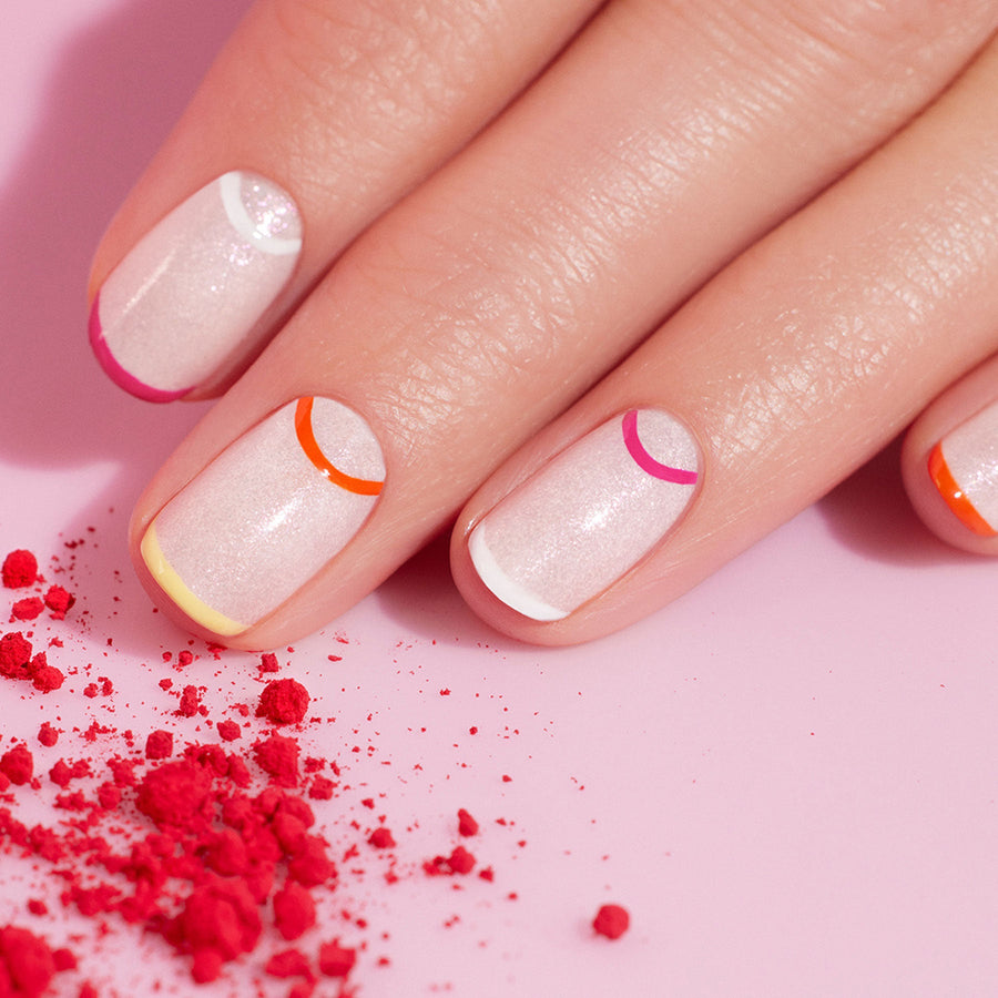 A hand with multicolored nails from Manucurist’s Smile! rests on pink and peach blocks, surrounded by four Smile! bottles and a Nail Art Brush in an inset circle—a bright, stylish setup perfect for summer nail art inspiration.