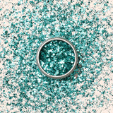 A small round container of Manucurist Turquoise Biodegradable Glitter rests on a plain light beige background, ready to enhance your nail art or gel polish designs.