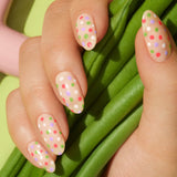 A hand with decorated nails holds a Manucurist Dot it better! near a box of five bottles, surrounded by flowers, a fish figurine, and a pink object. An inset highlights the dual-ended Dot it better! tool for creative nail art.