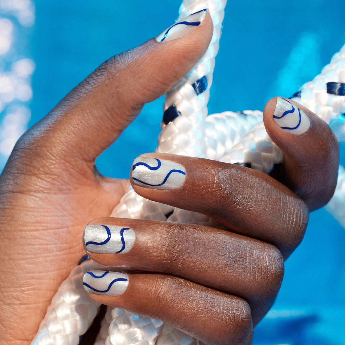 Two bottles of Manucurist Midnight Swim nail polish are shown with a wooden cuticle stick, in front of a coiled white rope with navy accents on a white background—ideal for creative nail art.