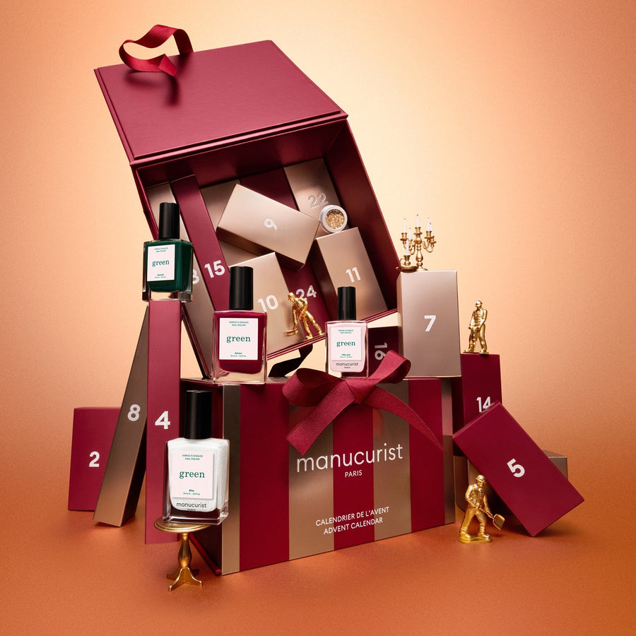 A red and gold Manucurist Advent Calendar Time to Shine! box with a ribbon, displayed on a peach background, featuring nail polishes, manicure accessories, and luxurious nail and hand care products like the innovative Active™ Shine Icy.