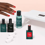 The Manucurist Discovery Kit features a white LED lamp, three nail polishes, a vitaminized base coat, remover, and four white nail clips—perfect for an ultra-glossy manicure—beautifully displayed on a light pink background.