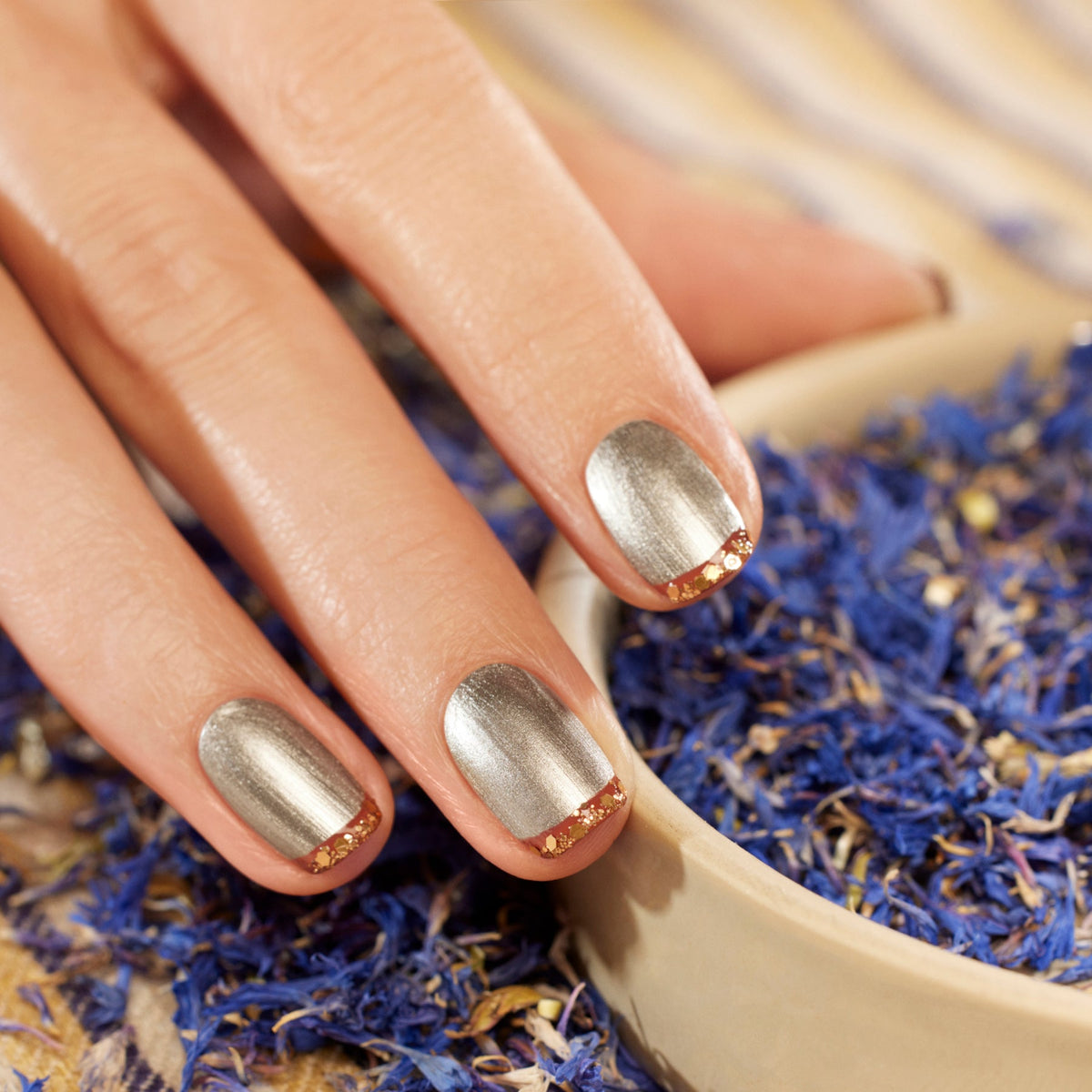 Two bottles of Manucurist Oriental Vibe nail polish and a pot of gold biodegradable glitter from the Autumn 2023 collection sit in a beige bowl on a striped cloth, surrounded by dried blue and purple flower petals. A manicure brush lies nearby.