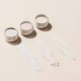 Three small tins of Manucurist Glitters and Effect Powders Trios in white, pink, and cream are open on a light surface, each with a swatch of their shimmery color brushed in front.