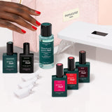 The Manucurist Starter Kit, featuring a pink Manucurist pouch, a sleek white LED nail lamp, green nail polish remover, four vibrant shades of nail polish, three white nail caps, and a dark green bottle—all arranged neatly on a light pink background—contains everything you need for an ultra-glossy manicure or to complete your gel polish kit.