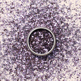 A small round container of Manucurist Lilac Biodegradable Glitter, featuring chunky, reflective mauve particles for nail art, sits on a light beige background and casts a soft shadow.