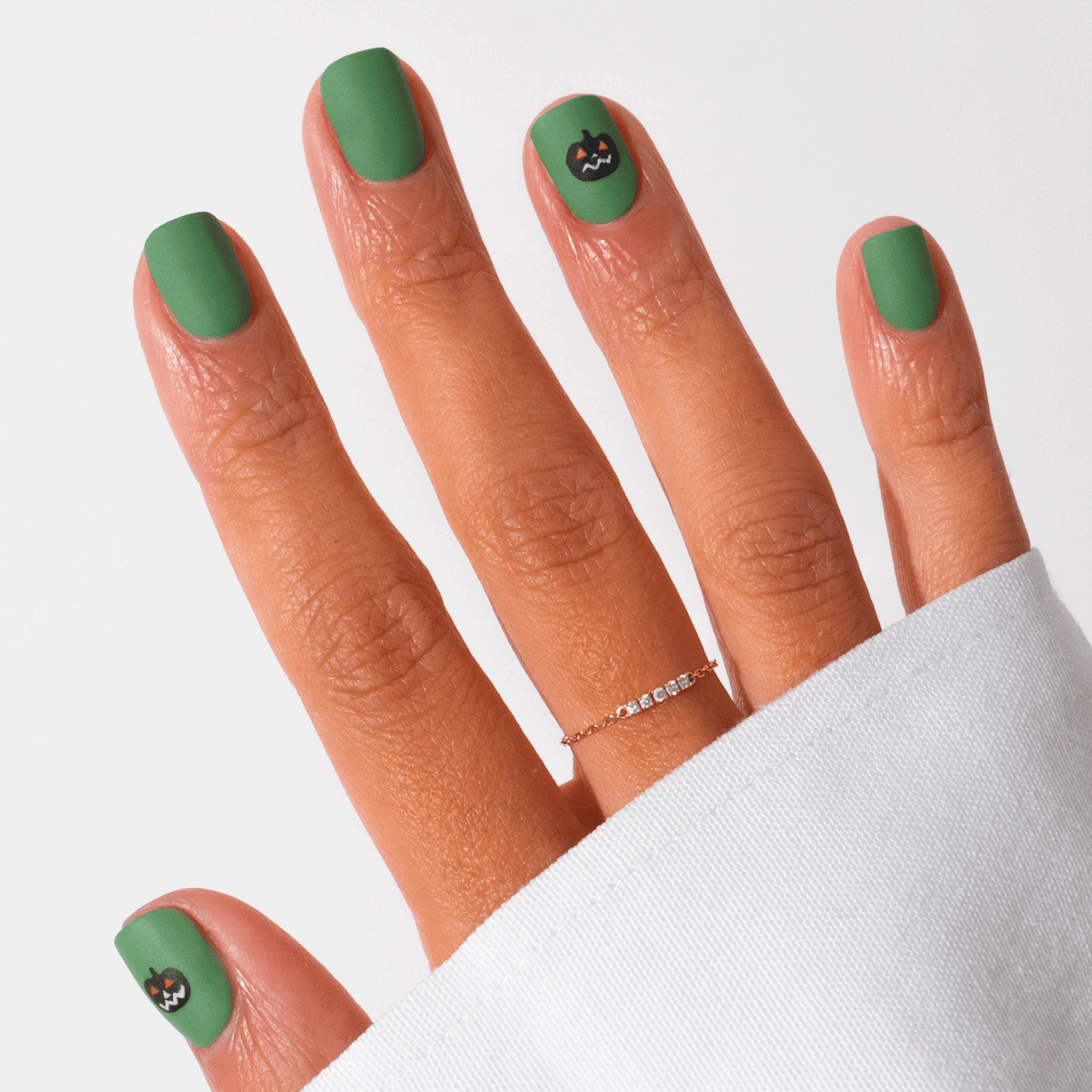Four bottles of Manucurist’s Pumpkin Patch nail polish in clear, orange, black, and green are displayed with a small open container of gold glitter on a purple and beige geometric background—ideal for Halloween nail art.