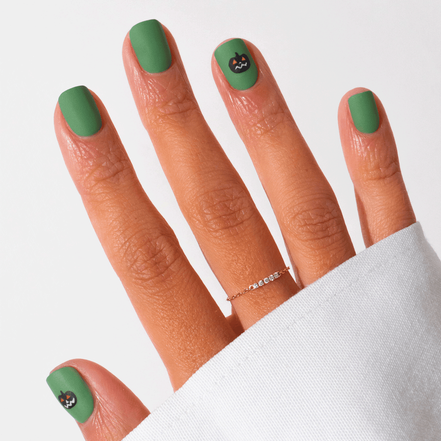 Four bottles of Manucurist’s Pumpkin Patch nail polish in clear, orange, black, and green are displayed with a small open container of gold glitter on a purple and beige geometric background—ideal for Halloween nail art.
