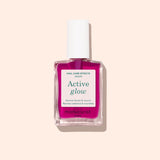 A bottle of Manucurist Active™ Glow Grape nail care polish, featuring a white cap and pink liquid, designed to enhance your natural shine, displayed against a light beige background.