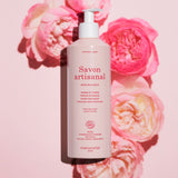 A pink bottle of Manucurist Organic Rose Hand  Body Wash - Cosmos Organic rests on a pink background with roses, its label noting a delicate rose scent and natural ingredients for luxurious hand and body care.