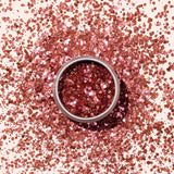 A small round container of Manucurist Pink Quartz Biodegradable Glitter, filled with shiny, chunky rose gold pieces, sits on a light beige background—perfect for adding eco-friendly sparkle to your nail art.