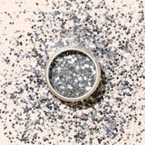 A round container of Manucurist Silver Biodegradable Glitter, designed for nail art, rests on a light beige background with a soft shadow cast to the left.